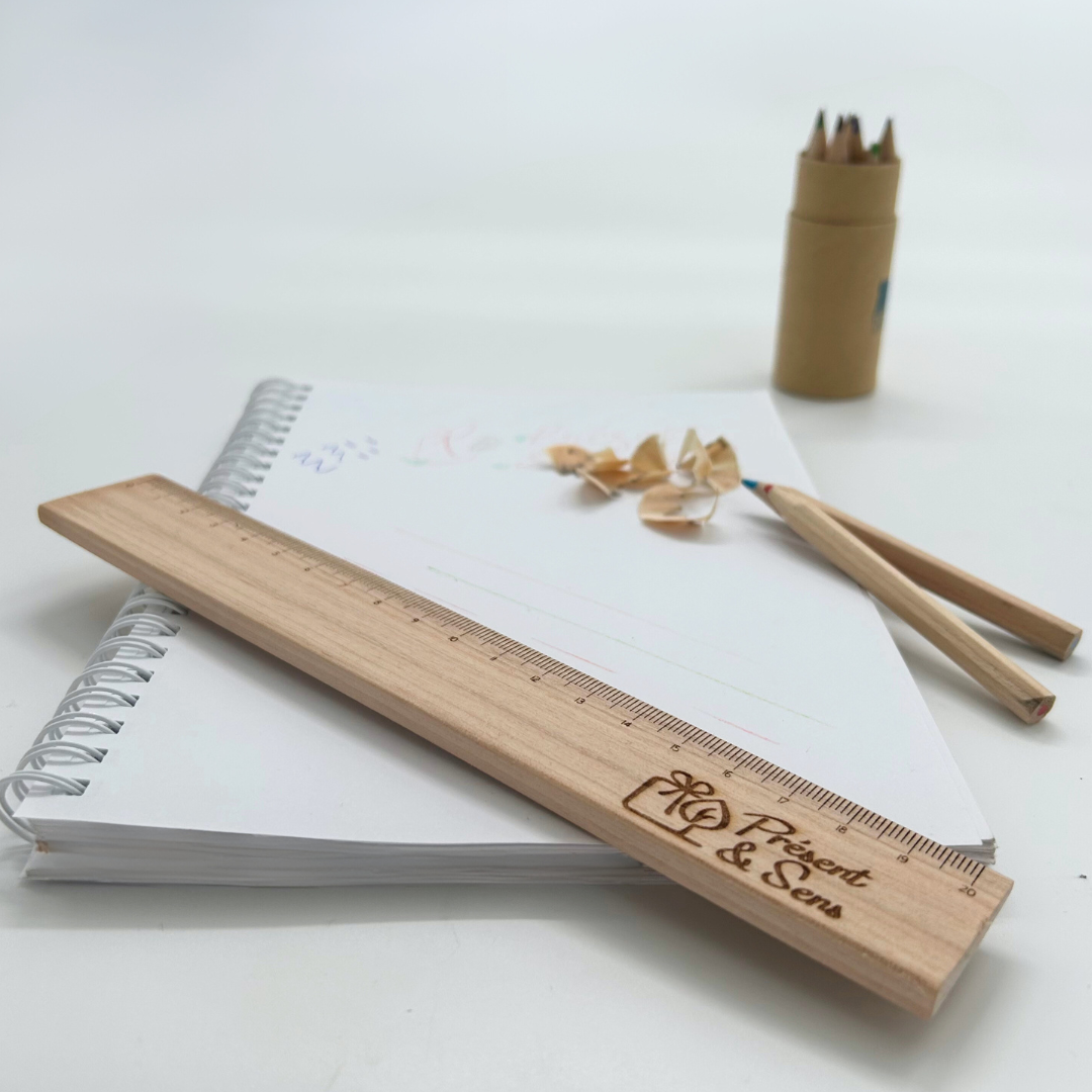 Règle en bois artisanale et gravée Une règle en bois, deux crayons de couleur et ses copeaux sont posés sur un cahier et à côté il y a une boîte de crayons de couleur.