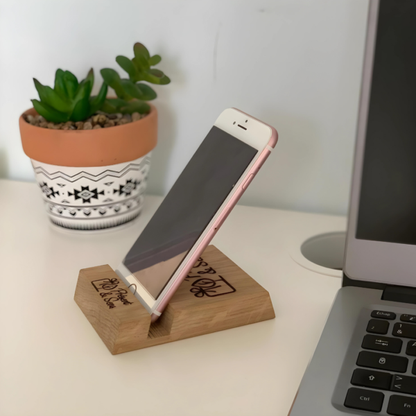 Support téléphone en bois gravé avec un mobile dessus, en arrière-plan un ordinateur et une plante.