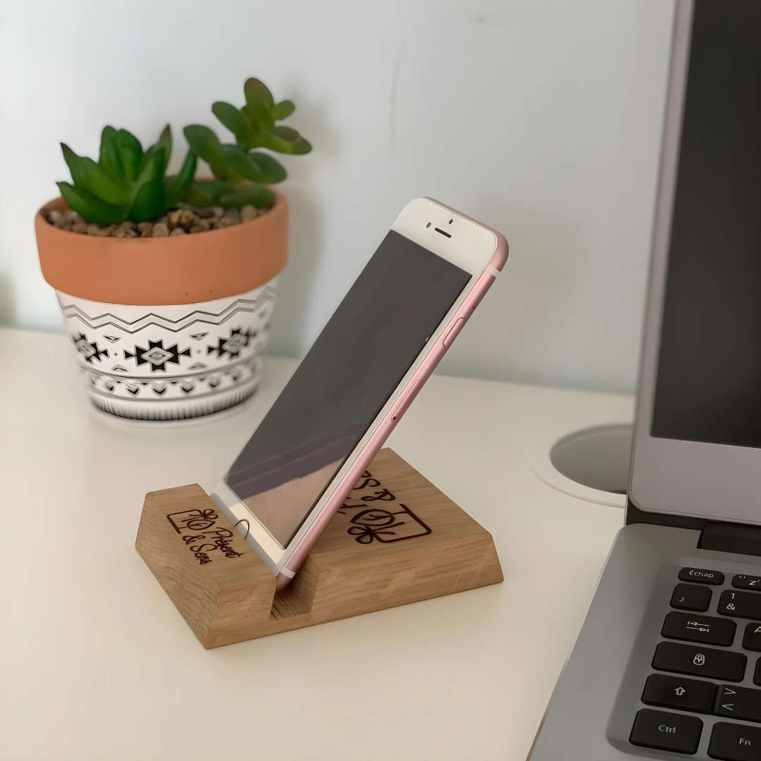 Support téléphone en bois gravé avec mobile Support téléphone en bois gravé avec un mobile dessus, en arrière-plan un ordinateur et une plante.