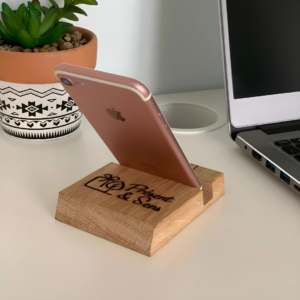 Dos du support téléphone en bois, gravé avec un portable posé dessus, en arrière-plan un ordinateur et une plante.