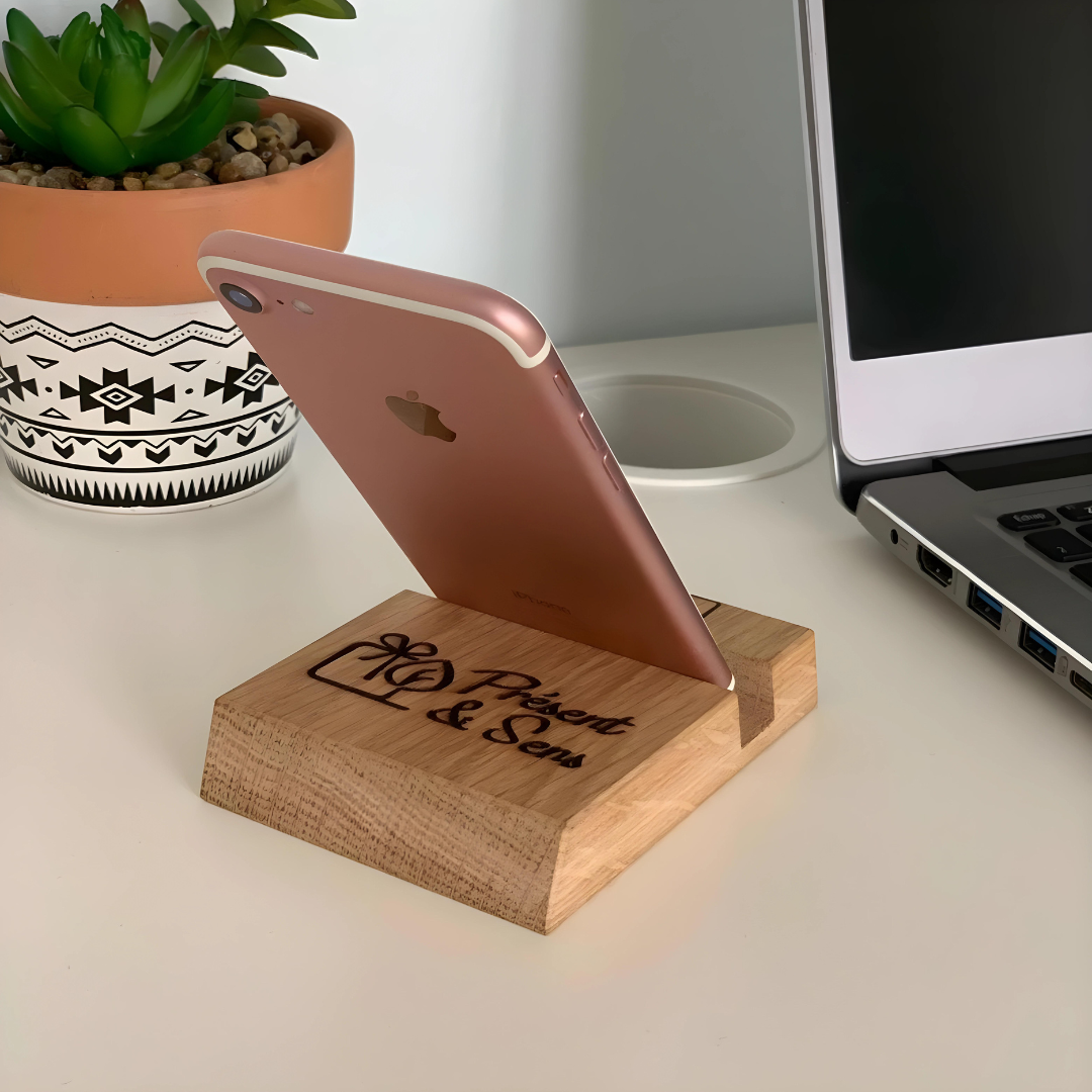 Support téléphone en bois gravé avec mobile côté dos Dos du support téléphone en bois, gravé avec un portable posé dessus, en arrière-plan un ordinateur et une plante.