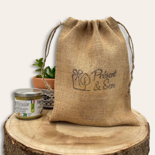 Pochon en toile de jute posé sur un tronc d'arbre coupé, un petit pot de plante sur l'arrière et un pot de tartine sur la côté.