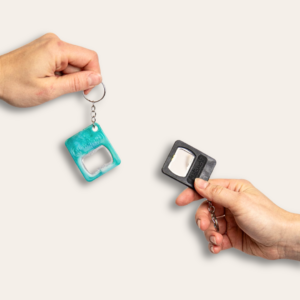 Deux mains tenant chacun un porte-clé décapsuleur en plastique recyclé noir et turquoise.