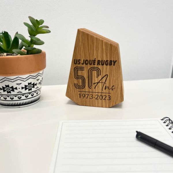 Trophée en bois gravé US Joué Rugby 50 ans et posé sur une table avec une plante et carnet à côté.