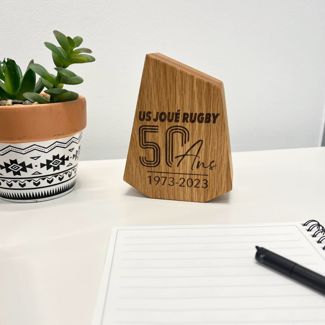 Trophée personnalisable Trophée en bois gravé US Joué Rugby 50 ans et posé sur une table avec une plante et carnet à côté.
